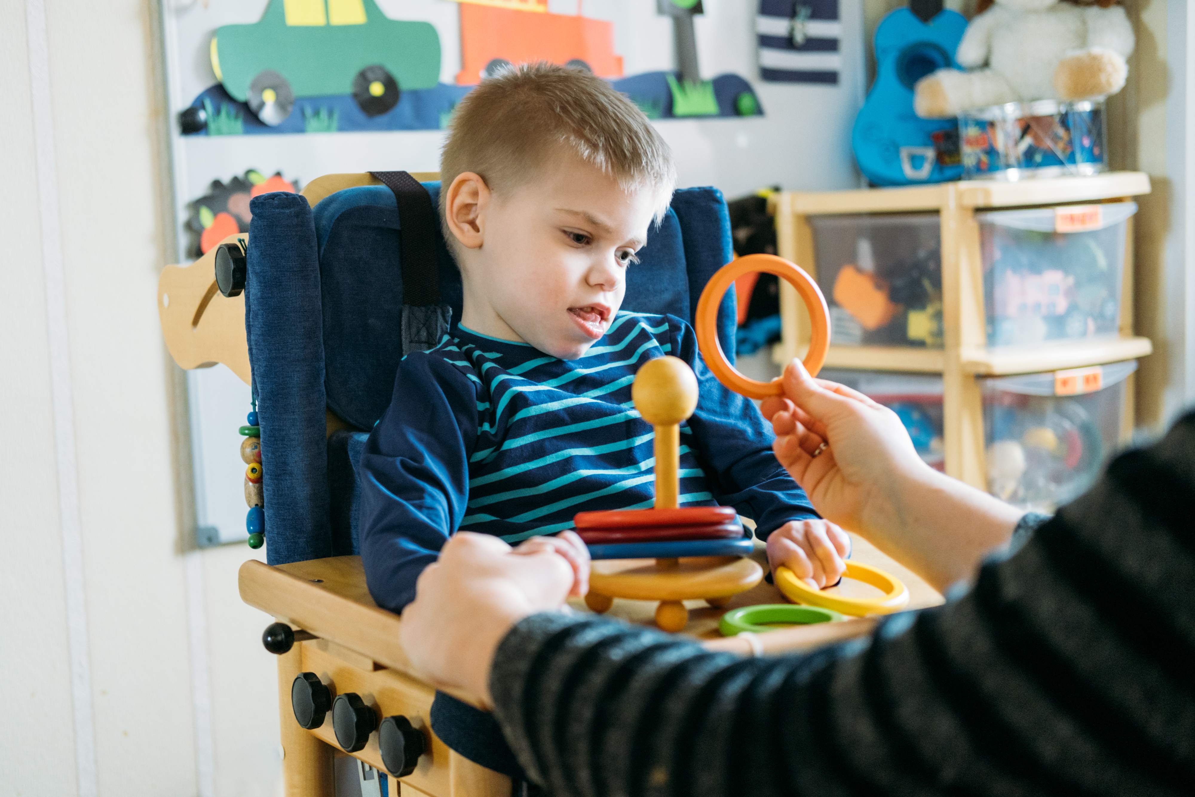 Young child in specialized therapeutic chair engaging with colorful developmental toys while receiving support from therapist