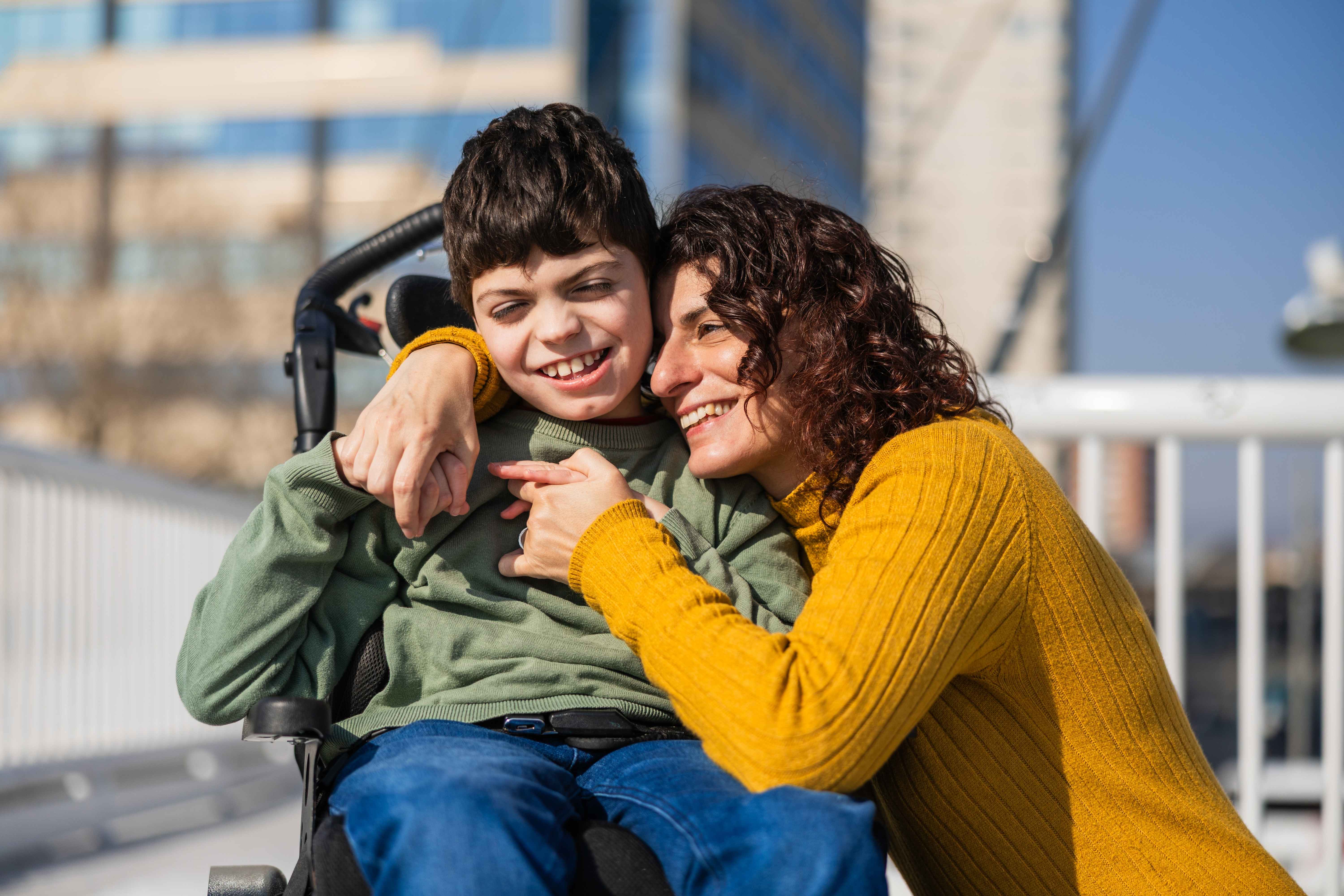 Loving moment between mother and child in wheelchair, both smiling warmly, representing family-centered support